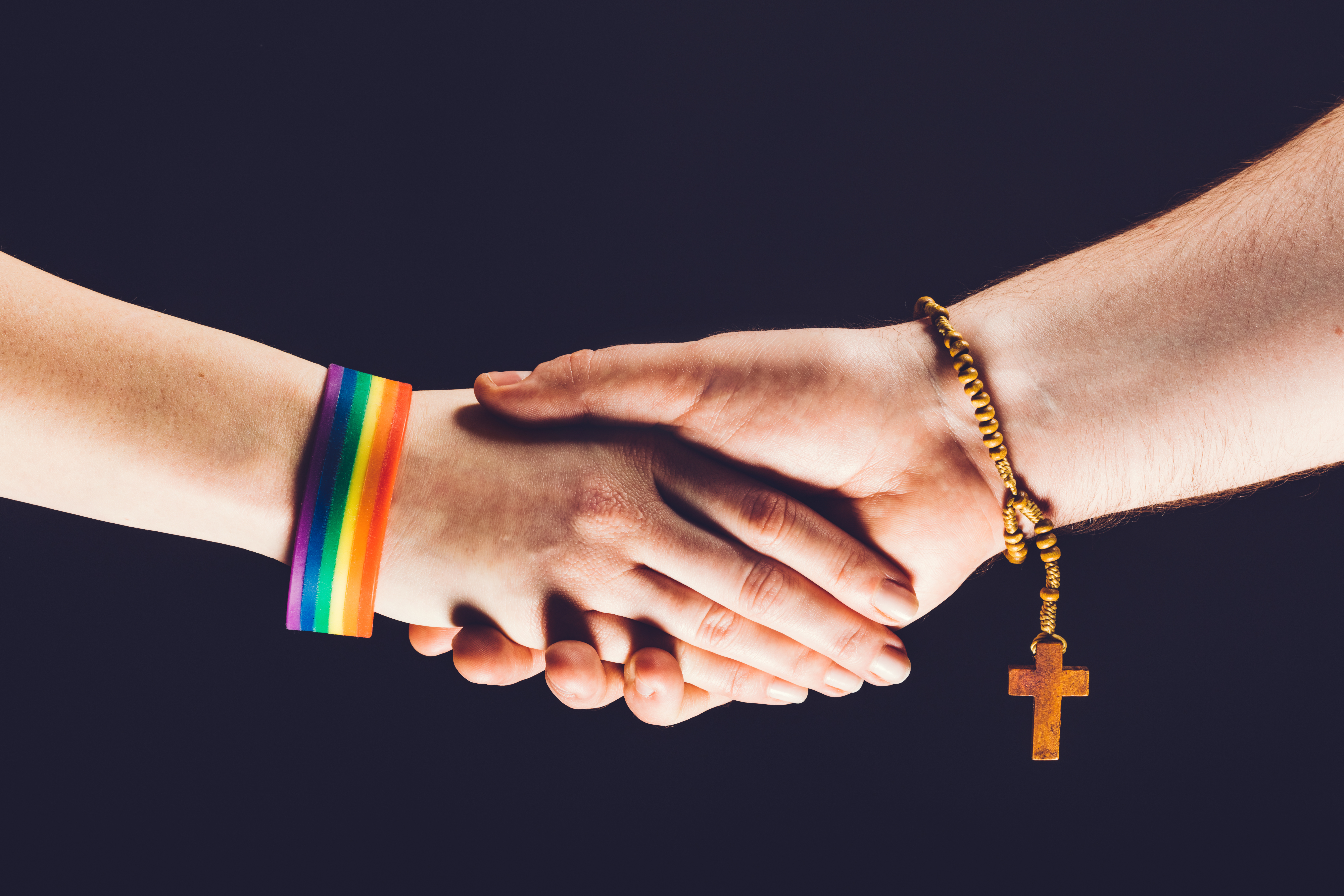Zwei Hände mit Regenbogenarmband und einer Gebetskette als Symbol für queeres Leben und Glauben.