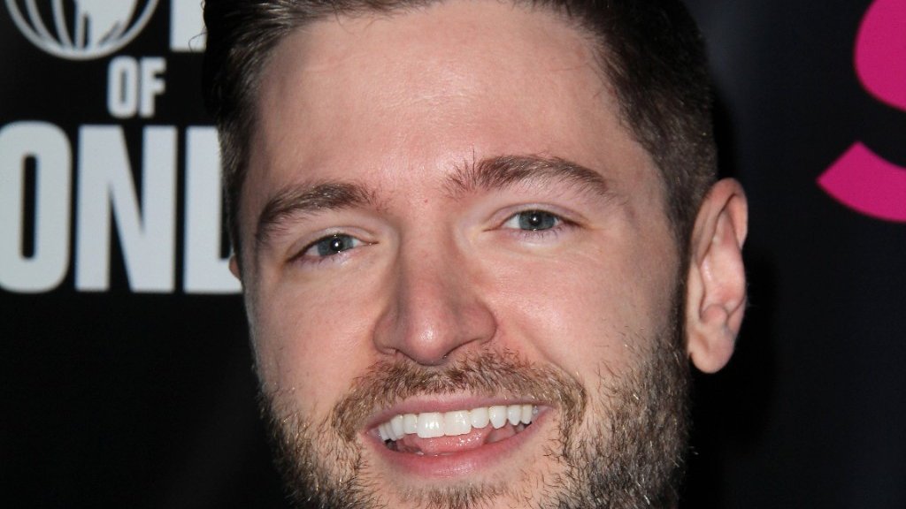 Lucian Piane lächelt in einem grauen Anzug mit schwarzer Fliege vor einer Veranstaltungswand.