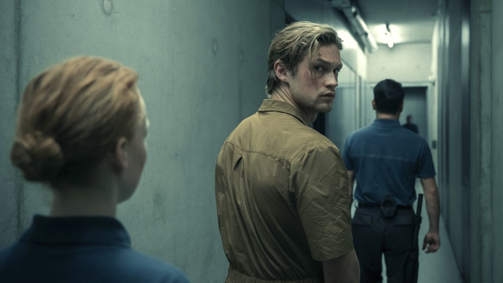 Szenenbild aus „Corpus Delicti“: Ein junger Mann in einem beigefarbenen Overall blickt in einem schmalen Gang über die Schulter zurück, während zwei uniformierte Personen ihn begleiten.