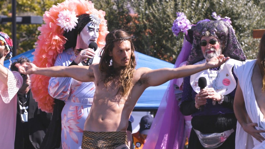 Filmstill aus Hunky Jesus: Jesus-Darsteller mit ausgebreiteten Armen zwischen den Sisters of Perpetual Indulgence