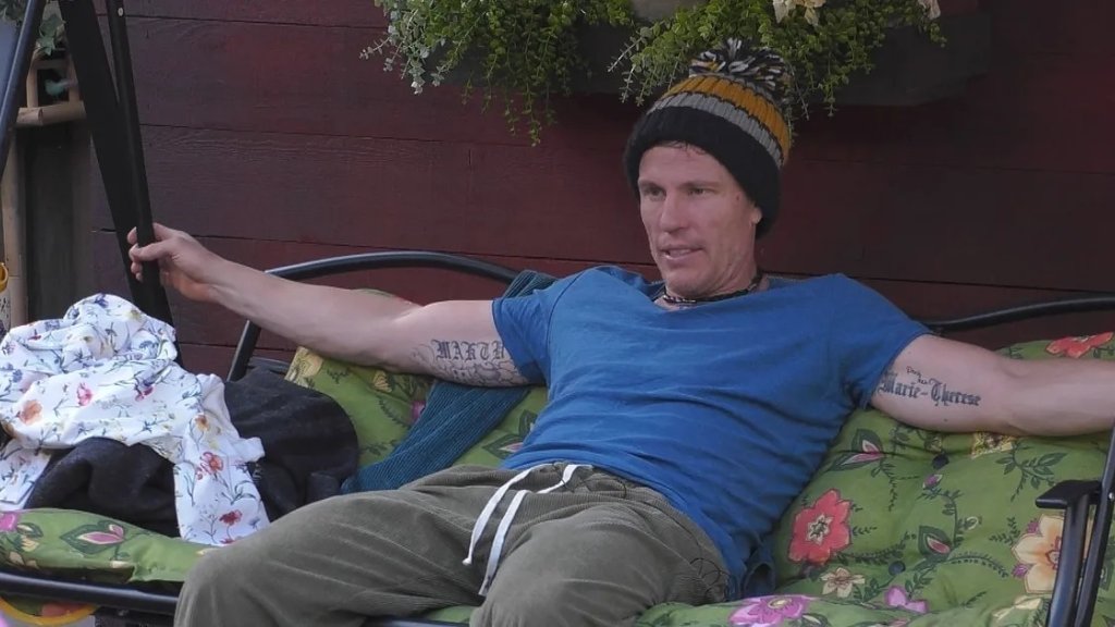 Christian Lell sitzt mit Mütze und blauem T-Shirt im Garten von Promi Big Brother.