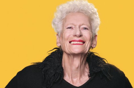Porträt der Künstlerin Dieter Rita Scholl mit kurzen weißen Haaren und schwarzer Kleidung vor leuchtend gelbem Hintergrund.