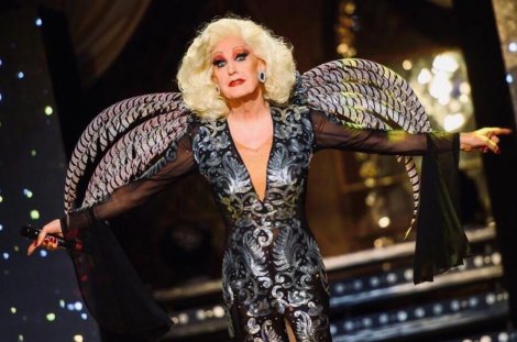 Drag Queen France Delon steht in einem schwarz-silbern verzierten Bühnenkostüm mit großen, flügelartigen Schulterelementen auf einer Bühne.