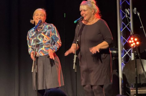  Zwei Darstellerinnen stehen bei einer Theateraufführung lächelnd an Mikrofonen auf einer beleuchteten Bühne.