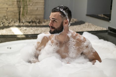 Bärtiger Mann sitzt mit nacktem Oberkörper in einer mit viel weißem Schaum gefüllten Badewanne und hat Schaum in den Haaren.