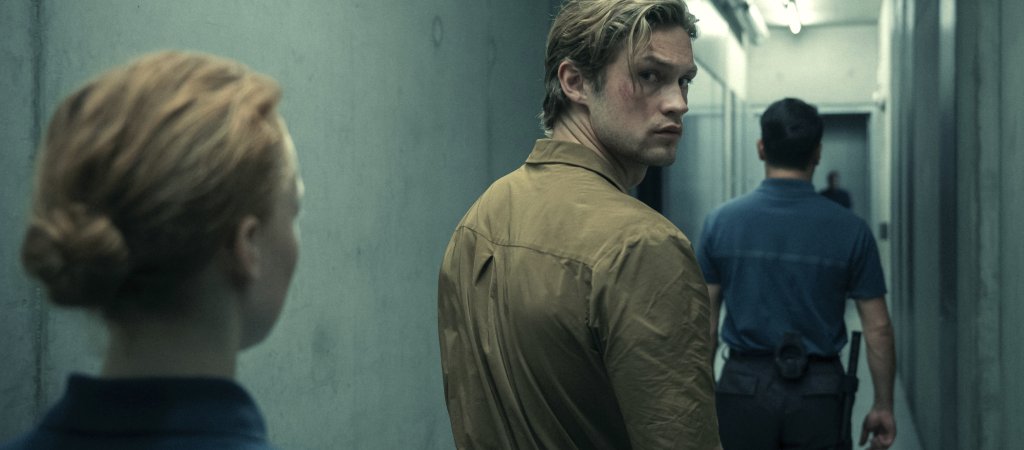 Szenenbild aus „Corpus Delicti“: Ein junger Mann in einem beigefarbenen Overall blickt in einem schmalen Gang über die Schulter zurück, während zwei uniformierte Personen ihn begleiten.