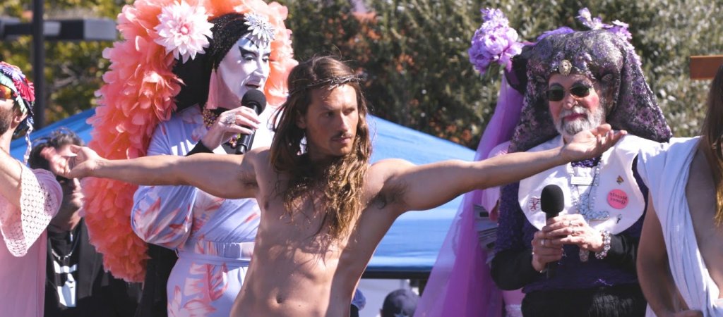 Filmstill aus Hunky Jesus: Jesus-Darsteller mit ausgebreiteten Armen zwischen den Sisters of Perpetual Indulgence