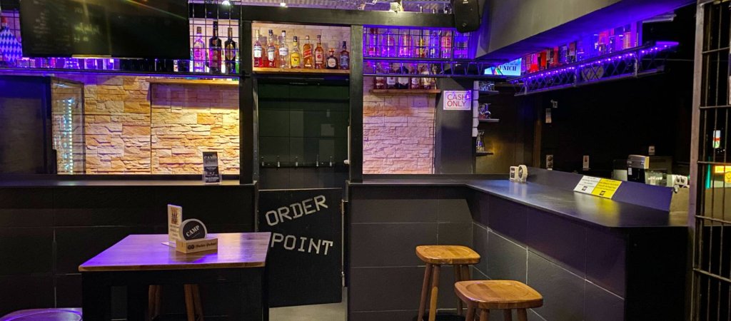 Barbereich des CAMP in München mit violett beleuchteten Regalen und Barhockern.