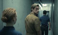 Szenenbild aus „Corpus Delicti“: Ein junger Mann in einem beigefarbenen Overall blickt in einem schmalen Gang über die Schulter zurück, während zwei uniformierte Personen ihn begleiten.