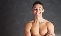 Lächelnder junger Mann mit nacktem Oberkörper und verschränkten Armen vor grauem Hintergrund.