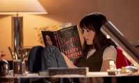 Szenenbild aus „Verity“: Lowen Ashleigh sitzt mit angezogenen Beinen in einem Sessel und liest konzentriert in einem mit zahlreichen Notizzetteln versehenen Buch.