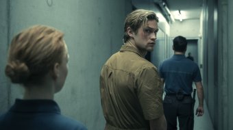 Szenenbild aus „Corpus Delicti“: Ein junger Mann in einem beigefarbenen Overall blickt in einem schmalen Gang über die Schulter zurück, während zwei uniformierte Personen ihn begleiten.
