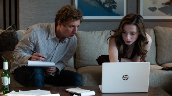 Szenenbild aus „The Social Reckoning“: Ein Mann mit Notizblock und eine Frau sitzen auf einem Sofa und betrachten gemeinsam einen geöffneten Laptop.