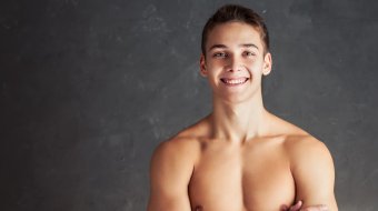 Lächelnder junger Mann mit nacktem Oberkörper und verschränkten Armen vor grauem Hintergrund.