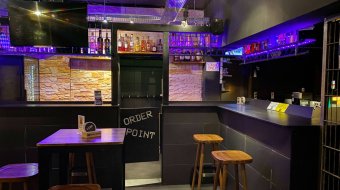 Barbereich des CAMP in München mit violett beleuchteten Regalen und Barhockern.