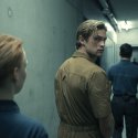 Szenenbild aus „Corpus Delicti“: Ein junger Mann in einem beigefarbenen Overall blickt in einem schmalen Gang über die Schulter zurück, während zwei uniformierte Personen ihn begleiten.