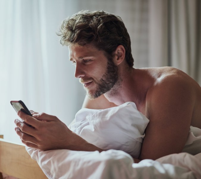 Symbolbild: Ein Mann sitzt im Bett und schaut auf sein Smartphone.