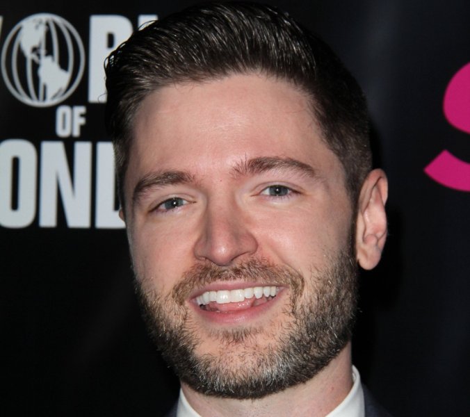 Lucian Piane lächelt in einem grauen Anzug mit schwarzer Fliege vor einer Veranstaltungswand.