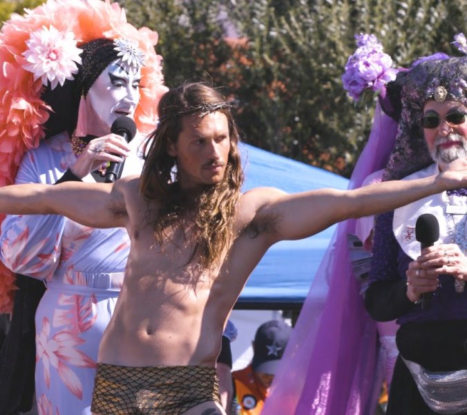 Filmstill aus Hunky Jesus: Jesus-Darsteller mit ausgebreiteten Armen zwischen den Sisters of Perpetual Indulgence